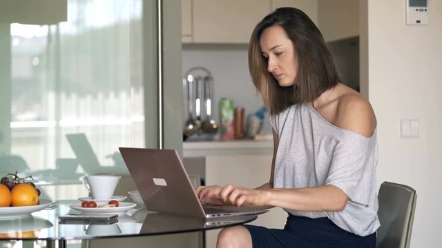 Young woman finishing working on laptop and drinking coffee in kitchen at home
