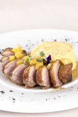 Luxurious dish with slices of duck breast, pineapple pieces, sweet sauce, with potato puree and parmesan, decorated with eatable flowers and red and green leafs, placed on white plate, light backgroun
