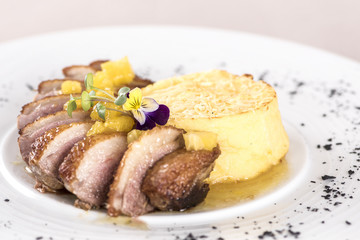 Luxurious dish with slices of duck breast, pineapple pieces, sweet sauce, with potato puree and parmesan, decorated with eatable flowers and red and green leafs, placed on white plate, light backgroun