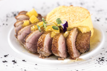 Luxurious dish with slices of duck breast, pineapple pieces, sweet sauce, with potato puree and parmesan, decorated with eatable flowers and red and green leafs, placed on white plate, light backgroun