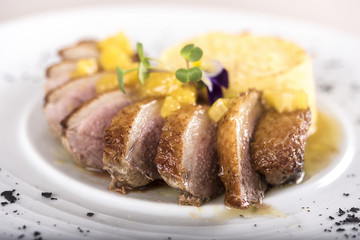 Luxurious dish with slices of duck breast, pineapple pieces, sweet sauce, with potato puree and parmesan, decorated with eatable flowers and red and green leafs, placed on white plate, light backgroun