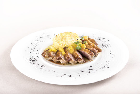 Luxurious dish with slices of duck breast, pineapple pieces, sweet sauce, with potato puree and parmesan, decorated with eatable flowers and red and green leafs, placed on white plate, light backgroun