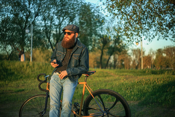 Bearded guy with mobile phone near fix bike. A brutal young man with a huge beard in sunglasses and a cap in the woods at sunset. Lifestyle.
