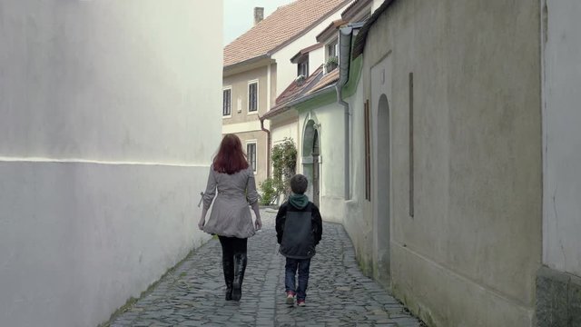 Mother and her son leave the camera in a narrow medieval aisle. A red-haired woman with a little boy is going to meet the sun. Positive, promising atmosphere. Static day stabilized shot.