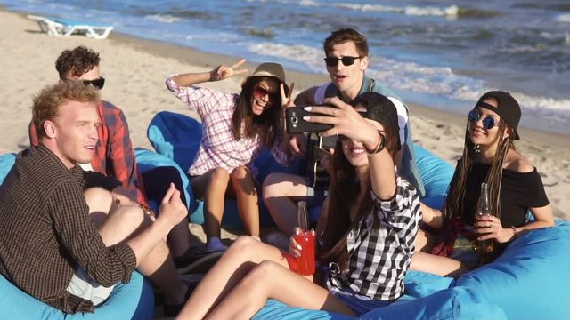 Young hipster girl recording a video or taking selfie of group of friends sitting on easychairs on the beach, playing guitar and singing on a summer evening. Slowmotion shot