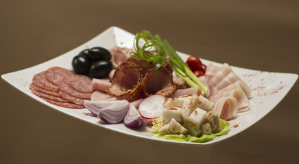 Cold cuts on plateau – different types of ham, bacon, salami, tomatoes, onion, green leafs, olives - white plate, brown background