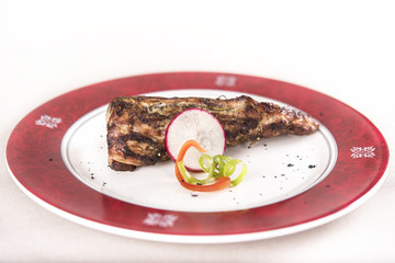 Grilled pork tenderloin, decorated with green and red leafs, placed on a white and red plate, light background
