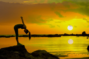 Asian woman playing yoga, exercise on rock at twilight light of evening at tropical beach with sunset and reflection of sun in the sea.