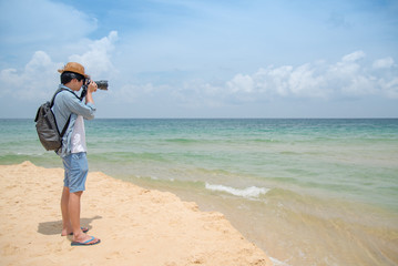 young Asian man photographer with jean shirt and hat taking photo of tropical island beach and turquoise sea, seascape background for summer holiday and vacation travel concepts