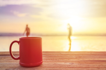 Red coffee cup on wood table at sunset or sunrise beach