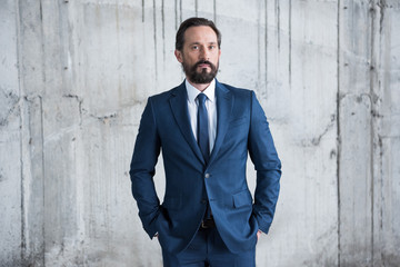 Confident middle aged businessman standing with hands in pockets and looking at camera