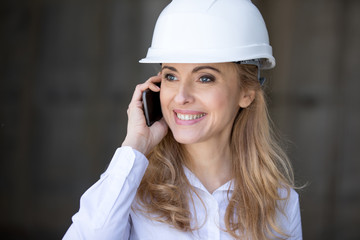 Smiling blonde businesswoman in hard hat talking on smartphone and looking away