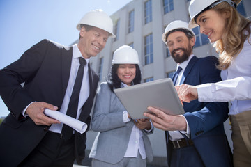 Smiling professional architects in hard hats holding blueprint and using digital tablet
