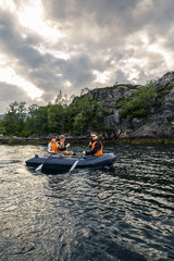 Group of young and attractive people in a boat in orange safe jackets is going somewhere by fjord. Exploring local places. Cloudy sky, Norway.