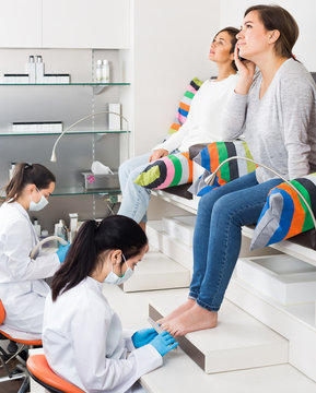 Woman Getting Pedicure By Professional In Beauty Salon And Using Phone