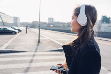Side view young woman walking in the city back light listening music headphones with smart phone hand hold - music, relaxing, enjoyment concept - obrazy, fototapety, plakaty