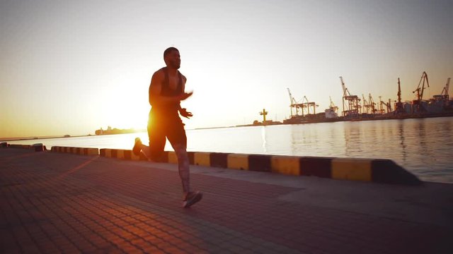 Strong diligent African American male jogging during sunrise along sea with port on background. Footage with sunflare in slwomotion
