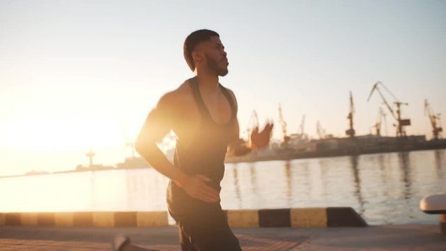 Young strong diligent African American male running along sea with port on background. Footage in slowmotion with sunflare