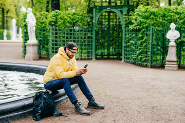 Outdoor shot of hipster guy with beard dressed stylish resting in park near fountain surfing social networks using his cell phone isolated over green park background. Sideways shot of serious man