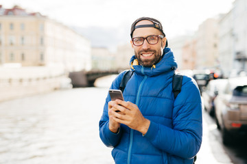 Happy man with beard wearing eyeglasses dressed in blue anorak holding backpack and mobile having happy look travelling in city watching sightseeings and making photos having exursion outdoor © VK Studio