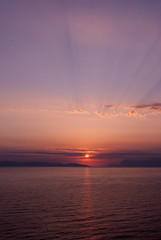 vertical image of sunset over the sea with purple blue hues and sunrays coming through clouds above
