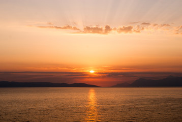 horizontal image of golden sunset with sun centrally low above the sea and sunrays coming through clouds above  