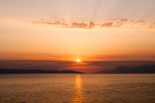 Fototapeta horizontal image of golden sunset with sun centrally low above the sea and sunrays coming through clouds above  