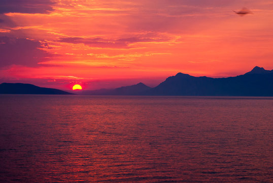 Fototapeta Horizontal image of deep coloured sunset with sea in red colour, black mountains on the horizon, and flying bird in a corner  