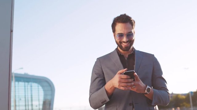 Rotation view of attractive man in a suit using his cellphone and happily smiling, looking around, gladly texting back in a bright sunlight. Modern lifestyle, social networks, active user.