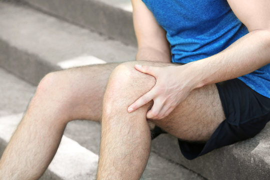 Young man suffering from pain in leg while sitting on steps outdoors, closeup