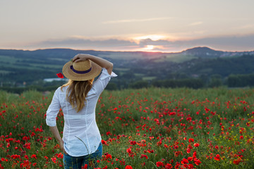 Young woman standing in poppy field dressed in jeans and white shirt watching sunset. Romantic portrait of a woman in straw hat on a meadow. 