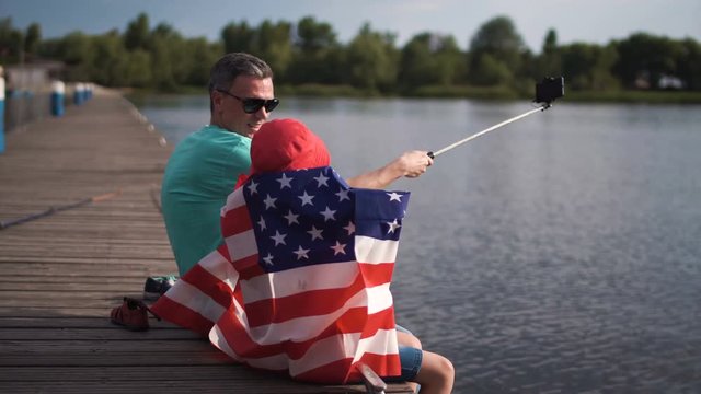 Son raise American flag while his dad make self picture using cell phone mounted on monopod or selfie stick