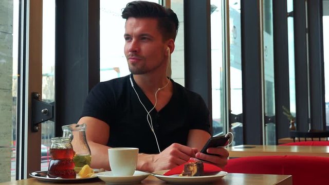 A young handsome man sits at a table with meal in a cafe and listens to music on a smartphone
