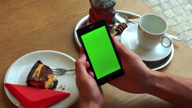 A man holds a smartphone with a green screen above a table with meal - closeup from above on the smartphone
