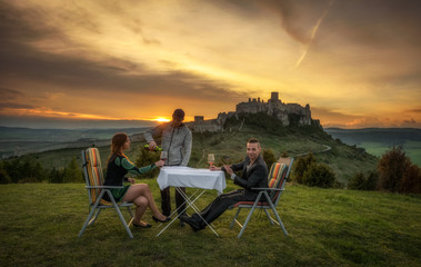Party of three people drink red wine in nature under the ruins of a castle at sunset.