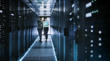 Female IT Technician and Male Server Engineer Talk and Discuss. They are in Working Data Center full of Rack Servers. Woman Holds Laptop. - obrazy, fototapety, plakaty