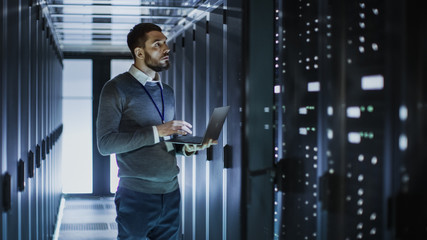 IT Technician Works on Laptop next to a Server Cabinet in Big Data Center. He Runs Diagnostics and...