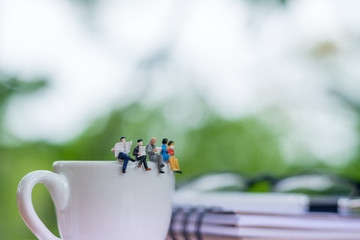 Miniature businessman reading book and sitting on the cup of coffee with glasses, notebook and soft focus green background using as a business concept.