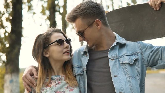 Young cute hipster couple walking and embracing along road with trees holding skateboards, youth freedom concept, 120FPS slowmotion