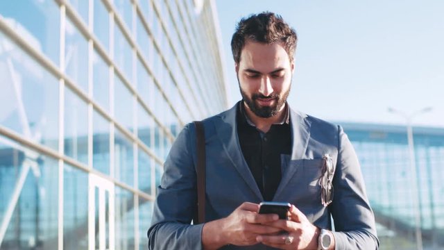 Self-confident handsome bearded man in fashionable suit walking by the airport entrance, using his cellphone and smiling to someone. Being happy, playful mood. Successful lifestyle.