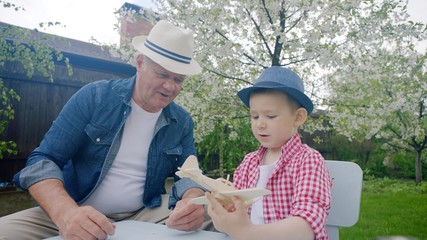 Granddad and his grandson are making wooden plane in the backyard on summertime