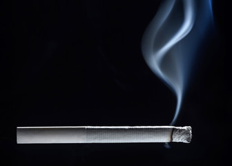 A white cigarette close-up begins to smolder tobacco and a trickle of smoke rises up against a dark background.