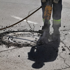 Worker at road demolishing asphalt with pneumatic jackhammer beside manhole
