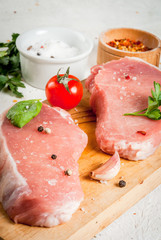 Raw pork, steak, cutlet. Meat, a source of protein. On a cutting board, on a white stone table. With spices, herbs and tomatoes for cooking. Copy space