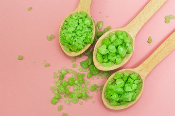 Green herbal bath salt in wooden spoons on a pink background.