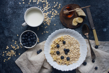 Organic oatmeal porridge with blueberry, banana, honey and milk on dark stone table, healthy lifestyle and diet concept, natural food, tasty breakfast, top view