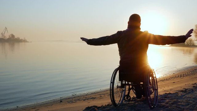 person on wheel chair, hands up, sundown, lonely disabled on wheelchair, male handicapped faith in future, self-confidence in human cripple, invalid outdoors, near river, back view