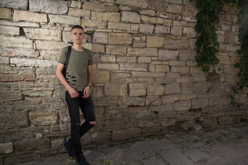 handsome man in glasses standing against a stone wall wearing a black t-shorts looking straight in to camera.