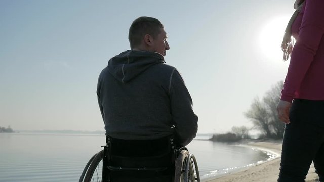 cripple person let hand to girlfriend, man handicapped and girl outdoors, back view, disabled near river on wheel chair, male on wheelchair, happy invalid pray for healing, sun glare