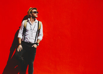 Young male model in hat white shirt and jeans hipster look opposite red background under hard light